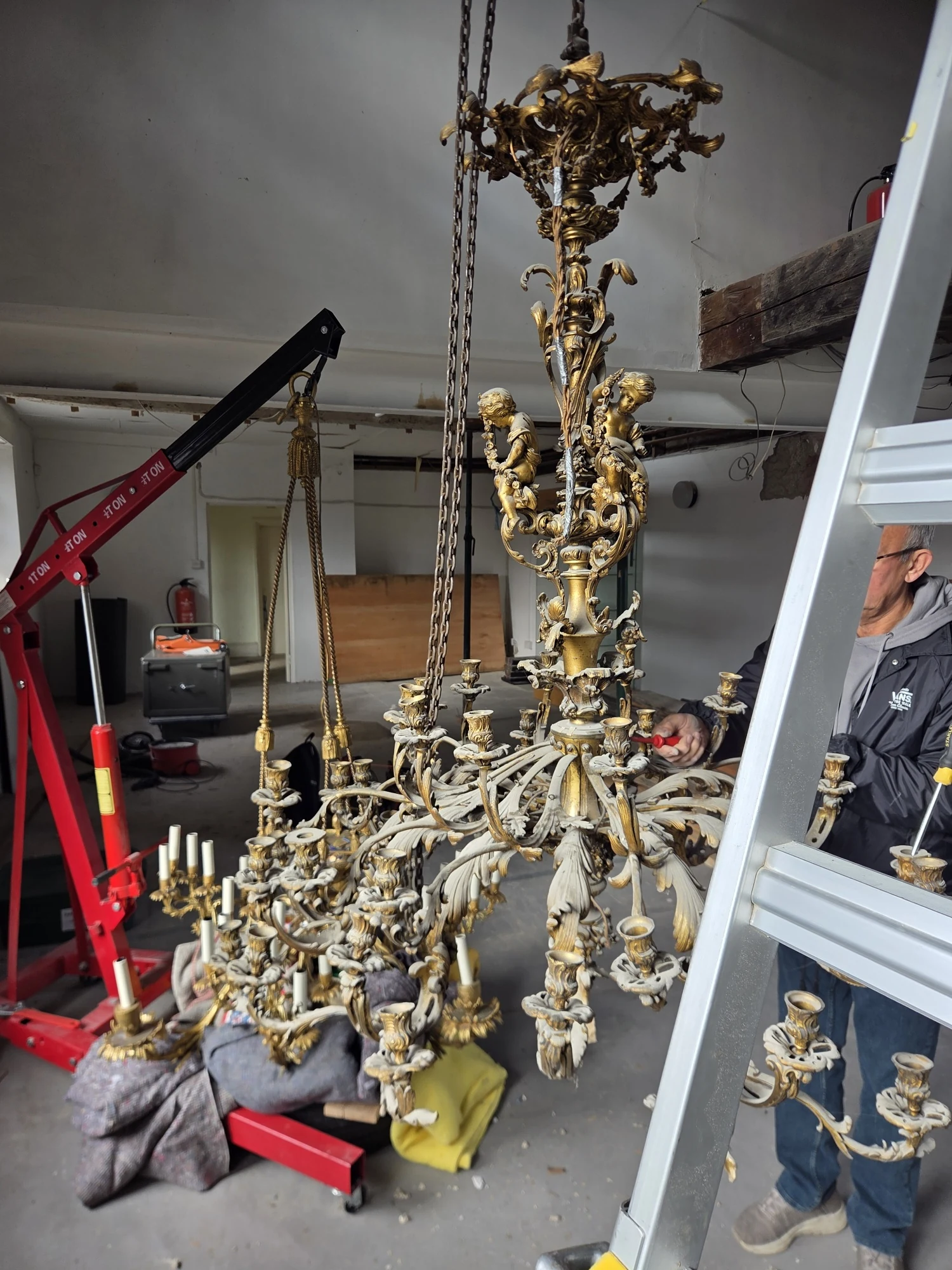The chandelier lowered within reach during dismantling, with ladders and red lifting equipment alongside.