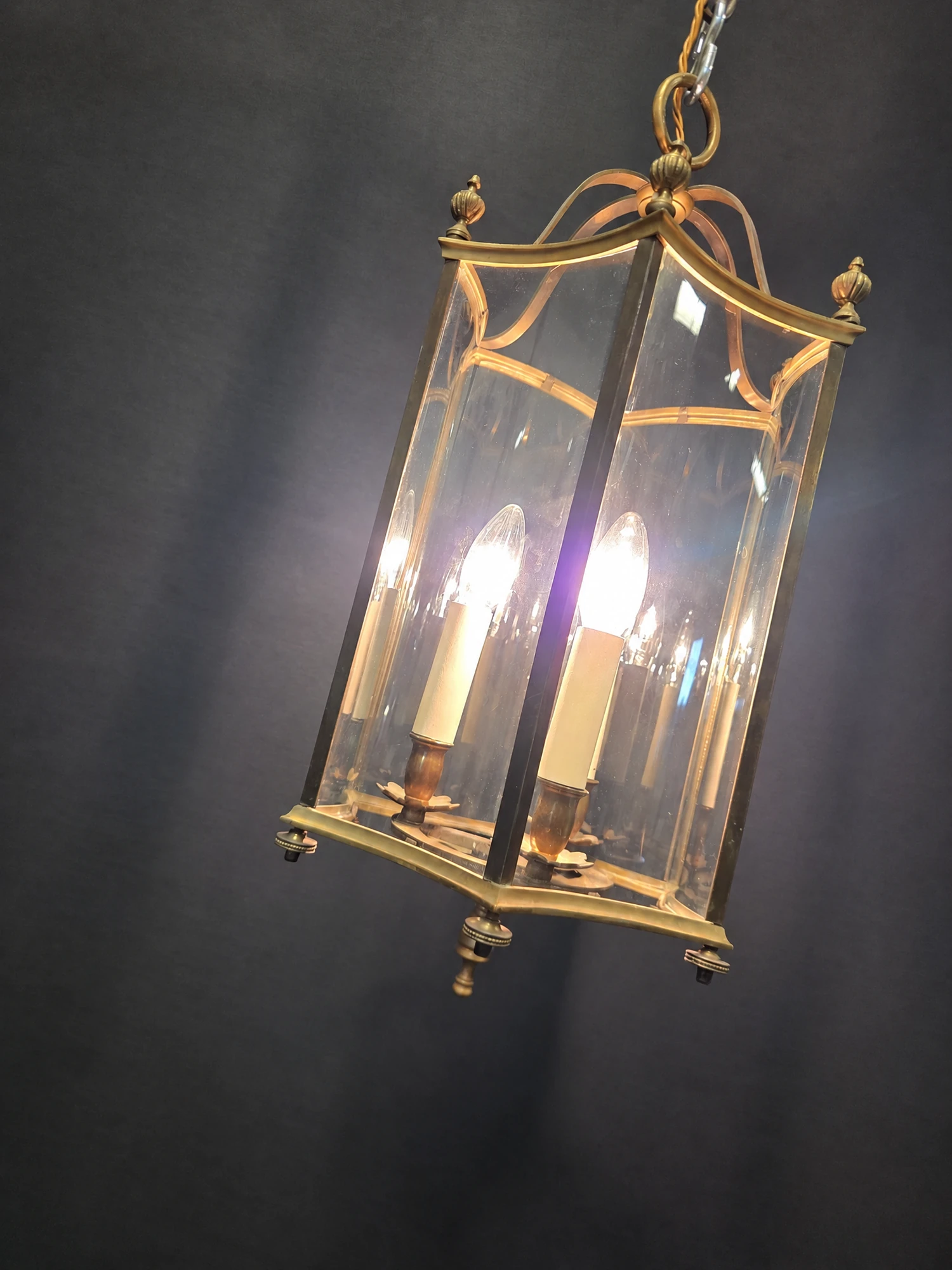 Illuminated hexagonal brass lantern viewed at an angle, showing the curved glass panels and scrolled brass top